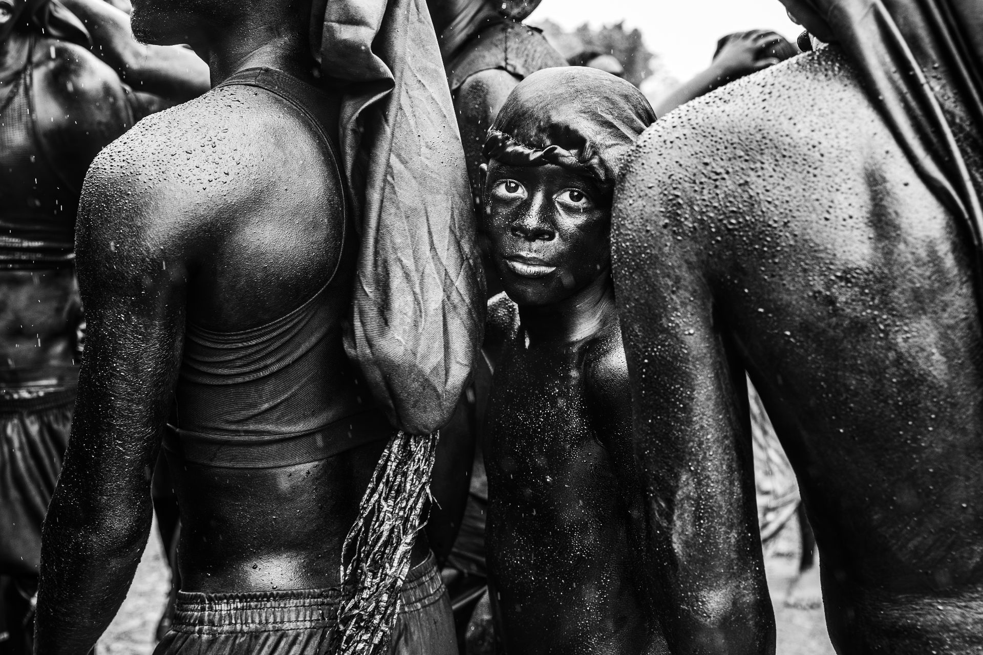 Black and white photo of shirtless, wet individuals standing close together, with one young person looking directly at the camera