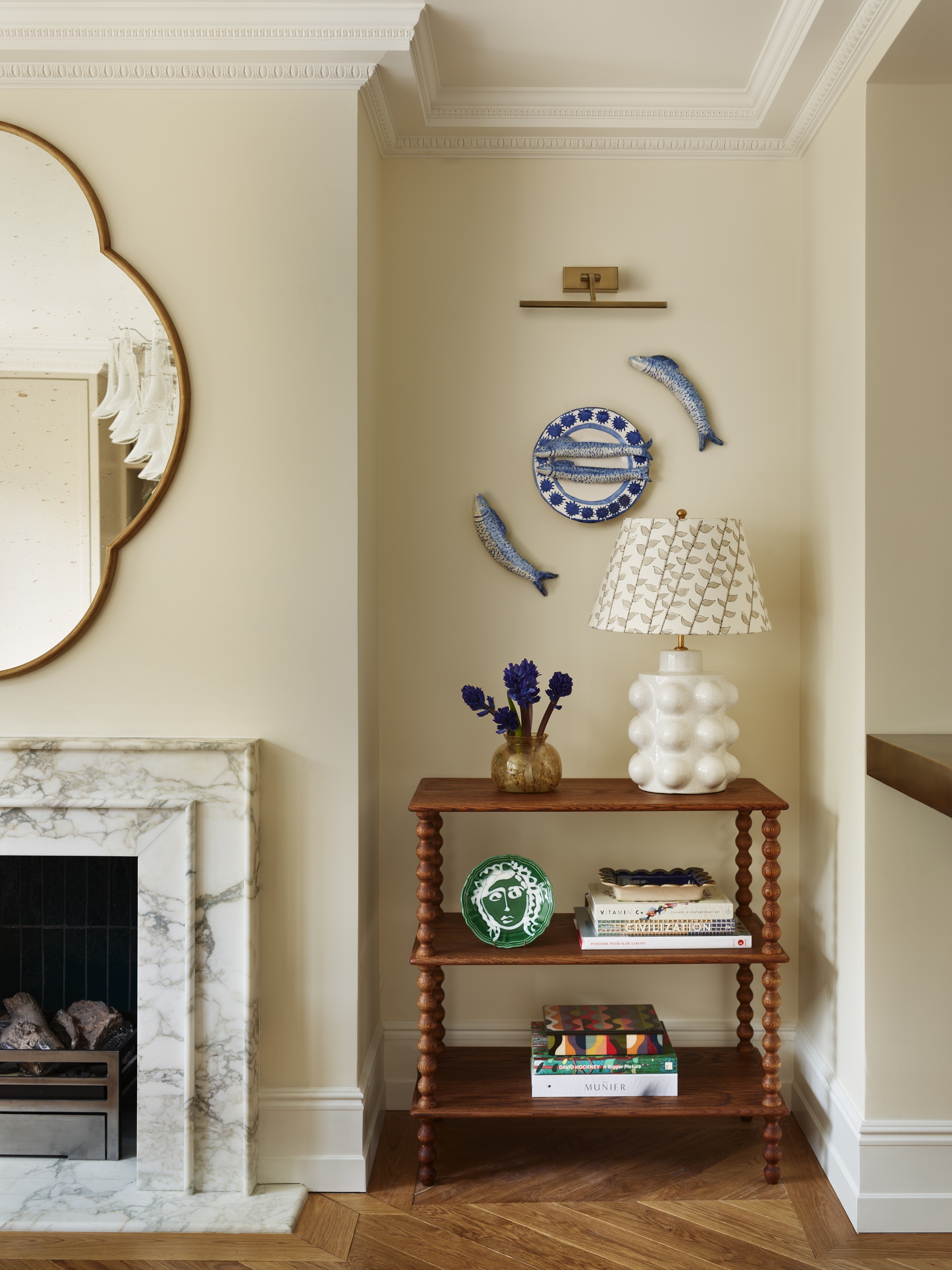 butter yellow living room with marble fireplace, gold mirror, bobbin table with lamp, and vases, and gallery wall with fish and plate and wall sconce