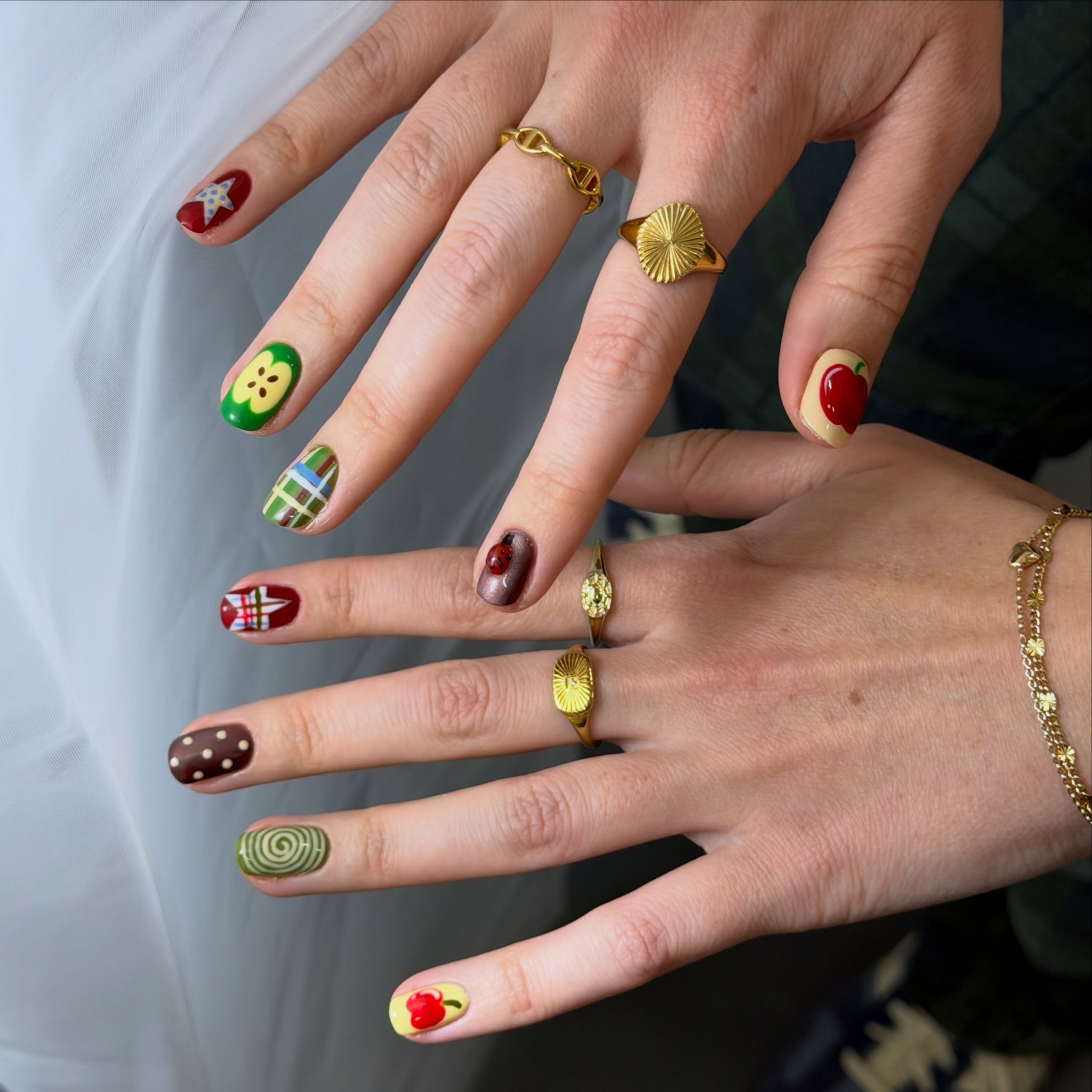 Short nails with colorful apple designs and gingham patterns, gold rings on fingers