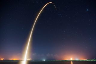 long exposure photo of rocket launch at night leaves a long trail of light through the starry sky.