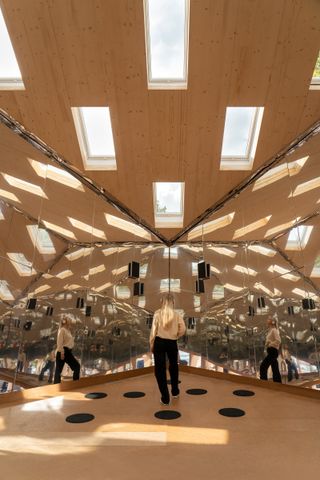 The interior of the Cube by Velux is seen through the trees. Wooden roof with skyline windows. Mirrors are on both walls that we see, with a woman standing in the center of the room.