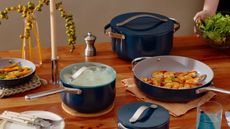 A set of dark blue ceramic non-stick pans on a wooden dining table. A sauce pan with a glass lid, a frying pan filled with fried shrimp, and a larger stock pot with a matching blue lid. 