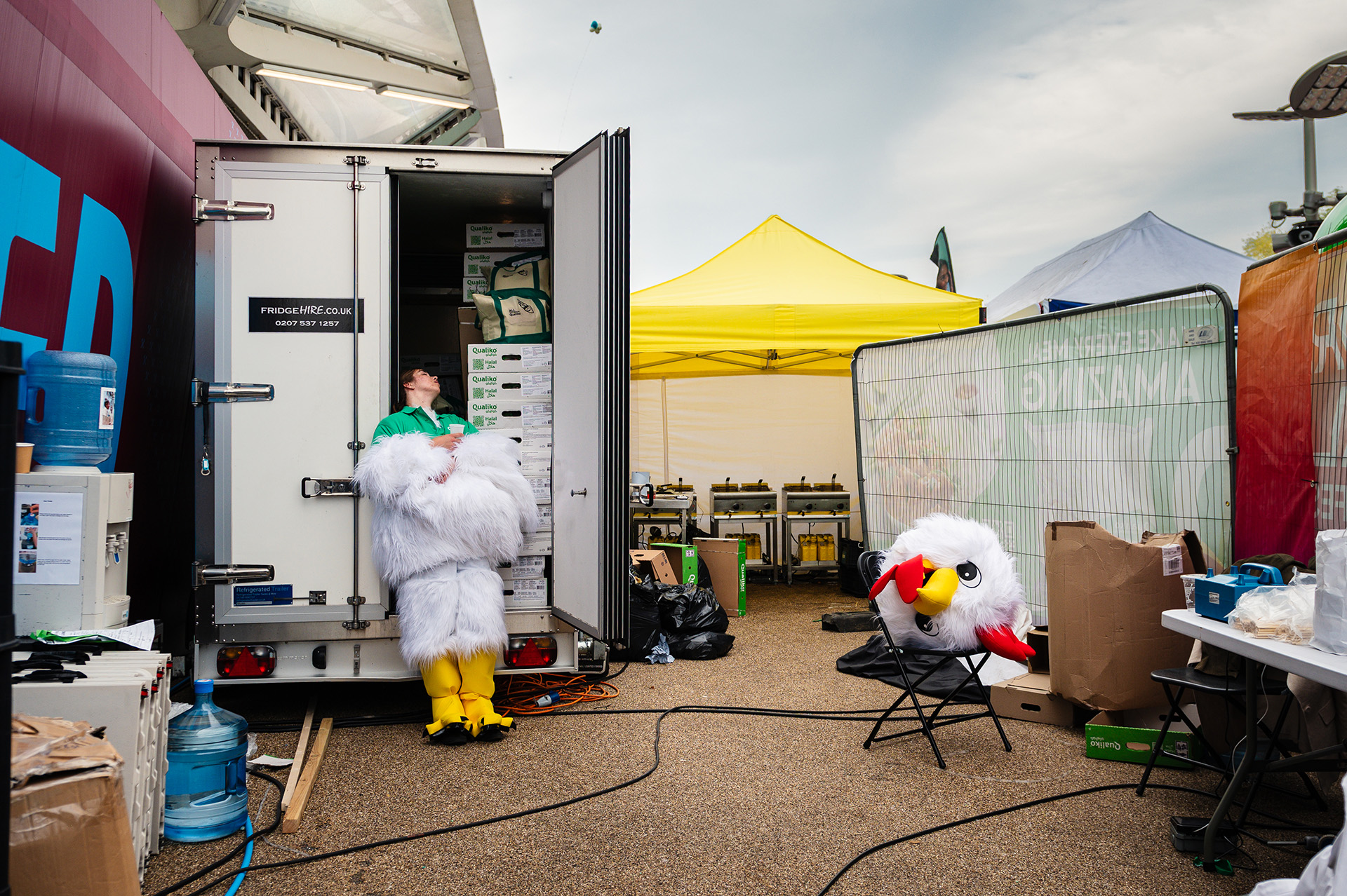 A performer at the World Halal Food Festival escapes the heat of their chicken mascot costume by cooling off in a refrigeration truck with a glass of water, costume head resting on a nearby chair.