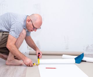 Man in blue polo shirt and khaki shorts is kneeling down on a wooden floor and measuring a strip of wallpaper