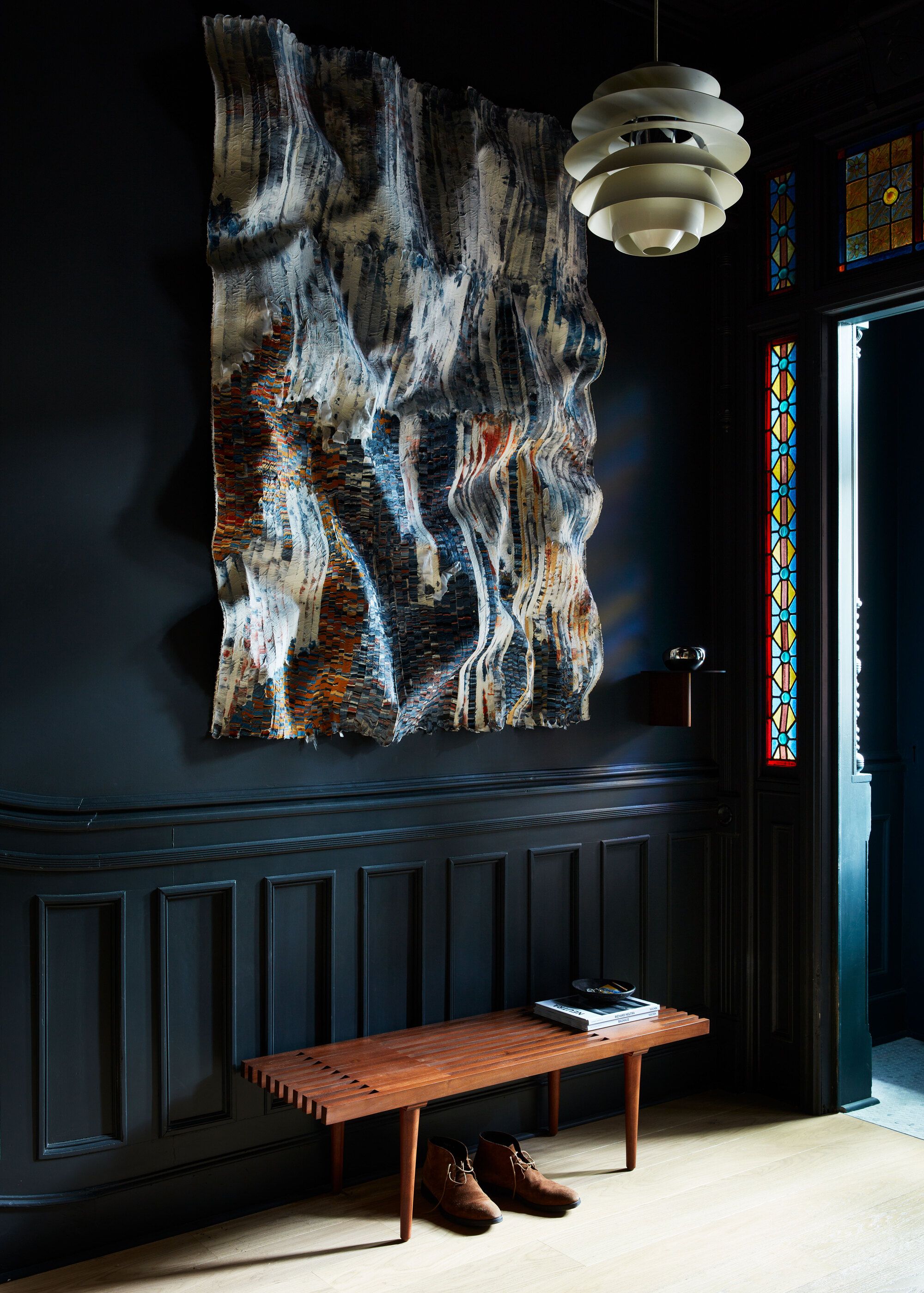 A dark blue entryway with a wooden bench, a large abstract artwork/scultpure on the wall in front of a pendant light, and original stained glass detailing in the door frame