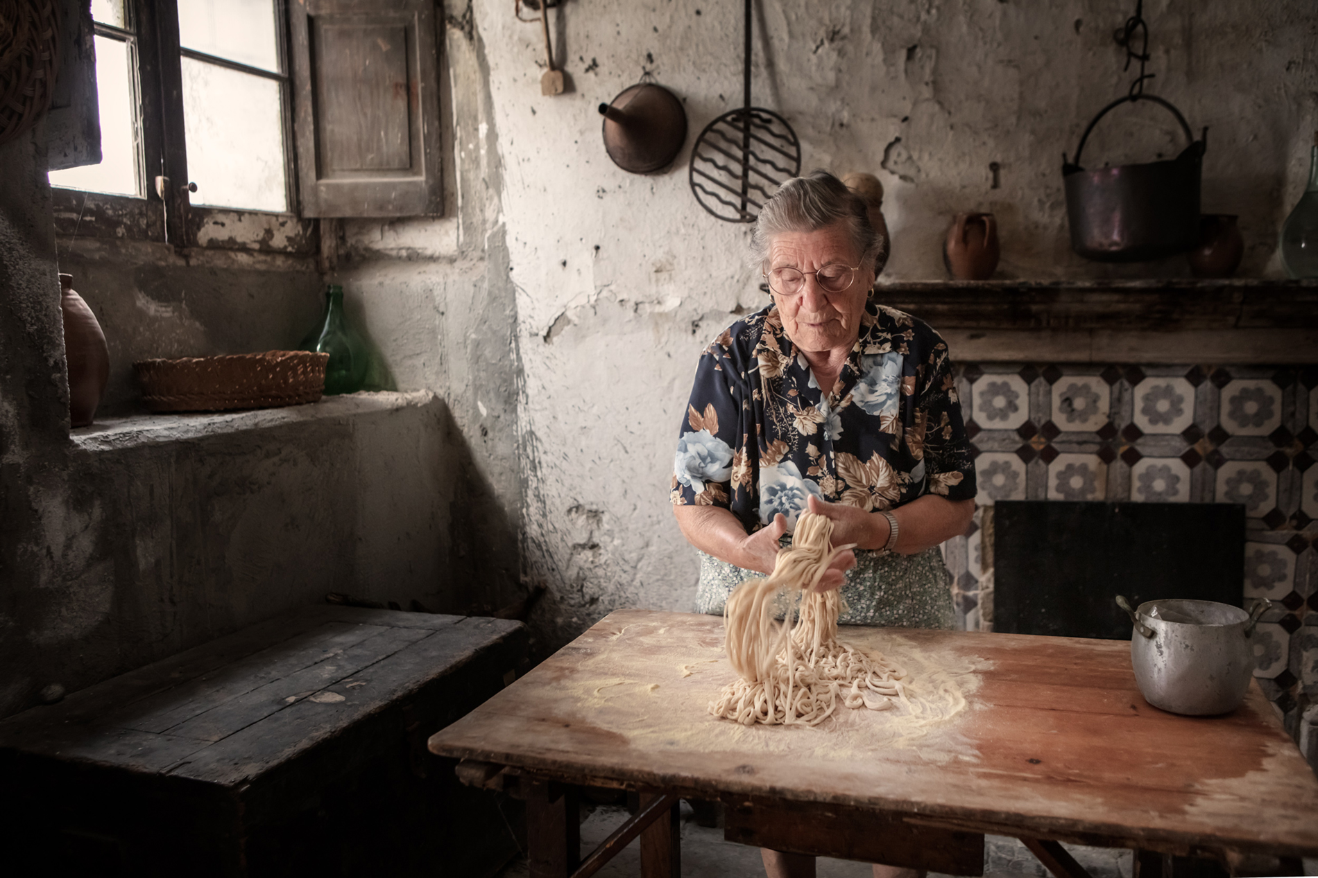 An elderly woman in a floral shirt kneads fresh pasta on a wooden table in a rustic kitchen with textured walls and vintage decor