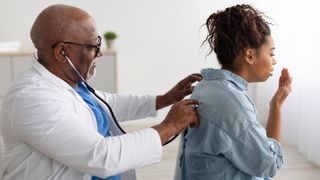Black male doctor checking breath of female patient using a stethoscope, listening to her lungs from the back. She is holding up a hand to cover her mouth and cough.