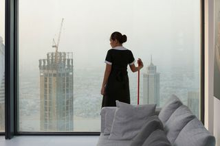 a woman standing behind a couch in front of a big window holding a broom