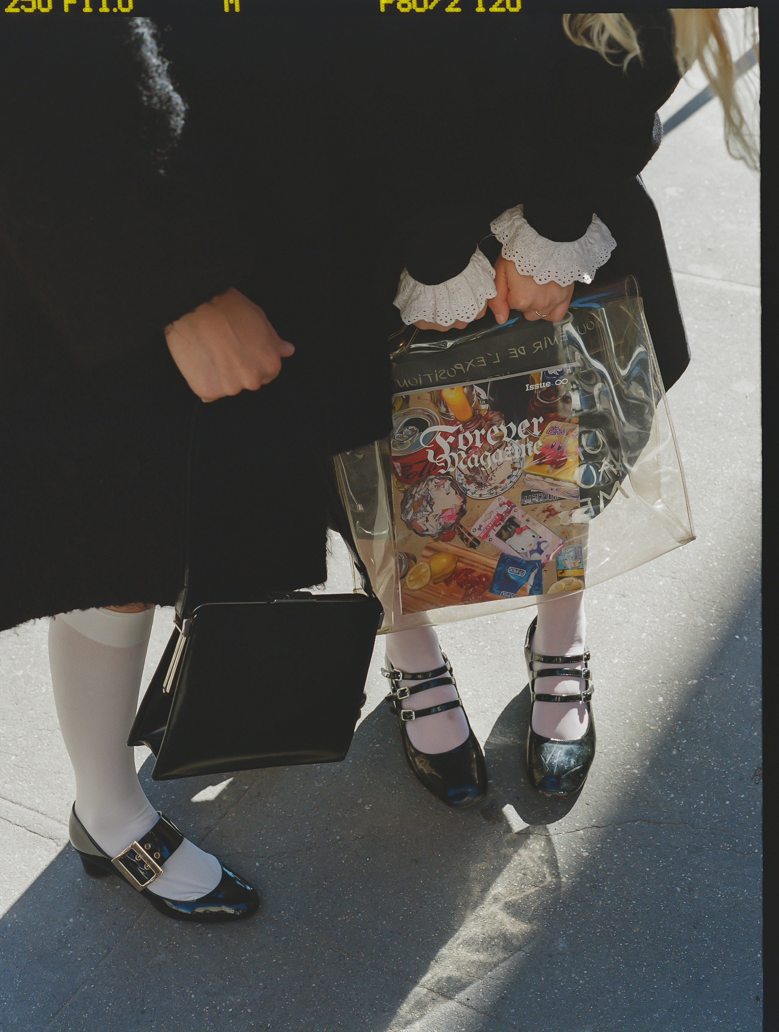 two women pose together from the waist down so you can see their black coats white socks and mary janes and the clear bag they're holding with the magazine forever in it
