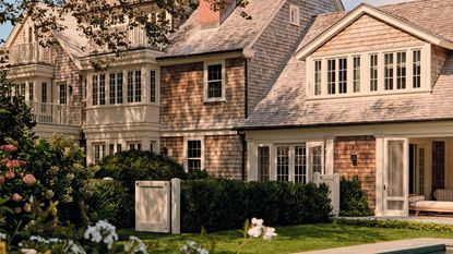 the exterior of a vast Hamptons brick home with soft beige exterior trims and a lush green pool yard