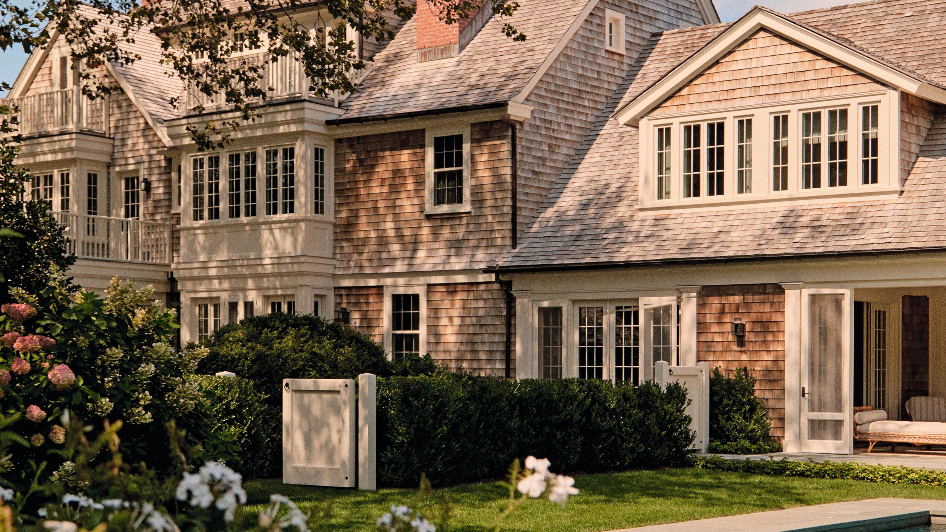 the exterior of a vast Hamptons brick home with soft beige exterior trims and a lush green pool yard