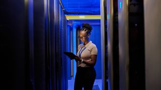 A young female engineer stood in a server room holding a tablet. Decorative: The engineer has medium dark skin and is lit in blue light.