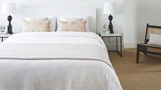 White bedroom with white walls, white bed with white bedding, jute carpet, two matching black lamps with white shades to either side of the bed, and a black and rattan chair with a white cushion.