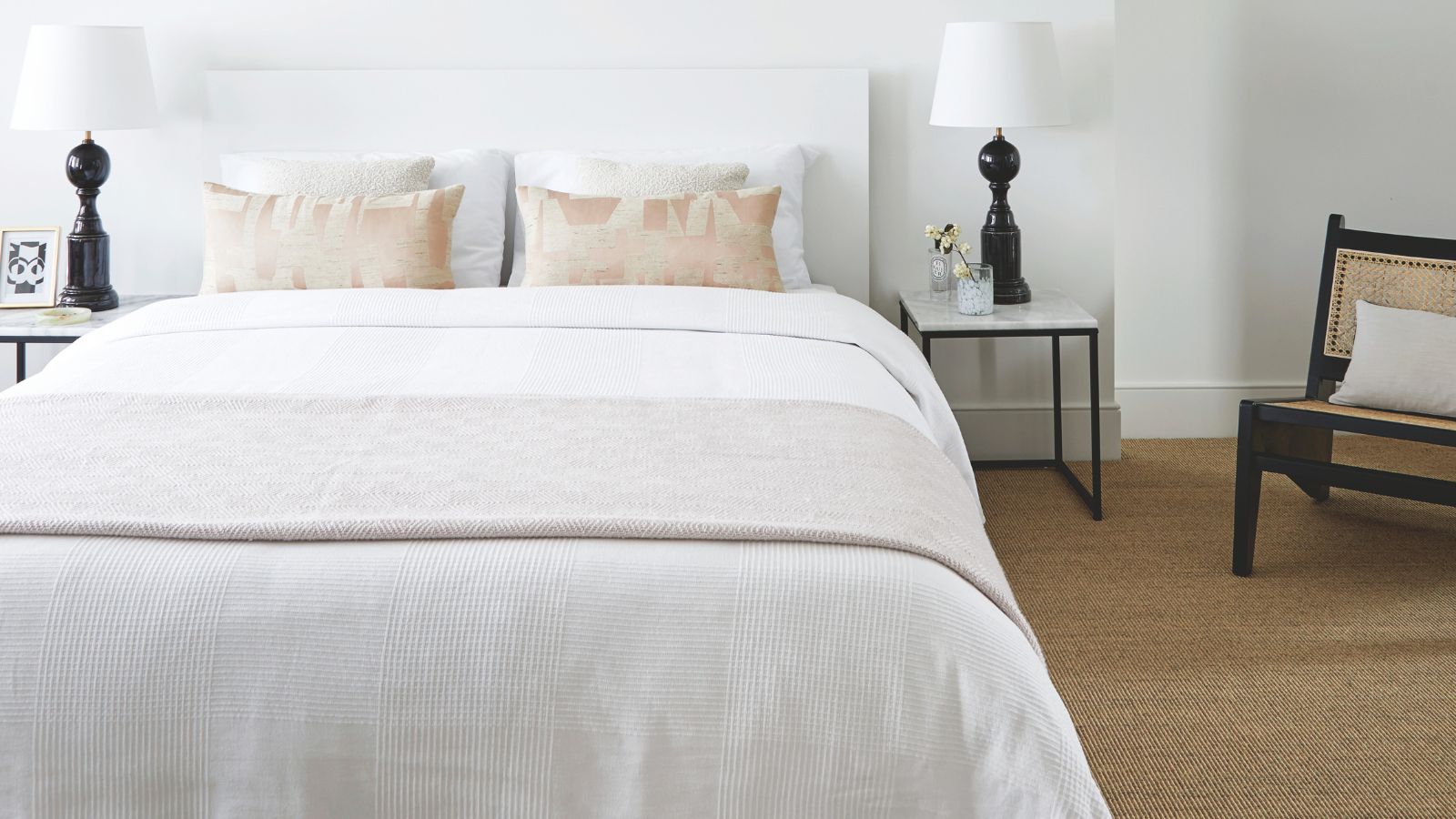 White bedroom with white walls, white bed with white bedding, jute carpet, two matching black lamps with white shades to either side of the bed, and a black and rattan chair with a white cushion.