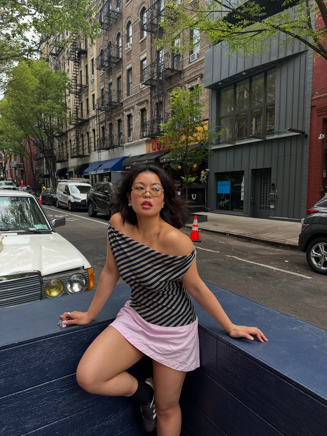 Fashion person Allison Ho wears the linen mini skirt trend in baby pink in a recent image. She has paired the skirt with a grey and black striped of the shoulder t-shirt, black and white Adidas sambas and clear oval glasses. She is leaning both arms on a dark blue decking and is posed towards the camera in a recent image on her Instagram.