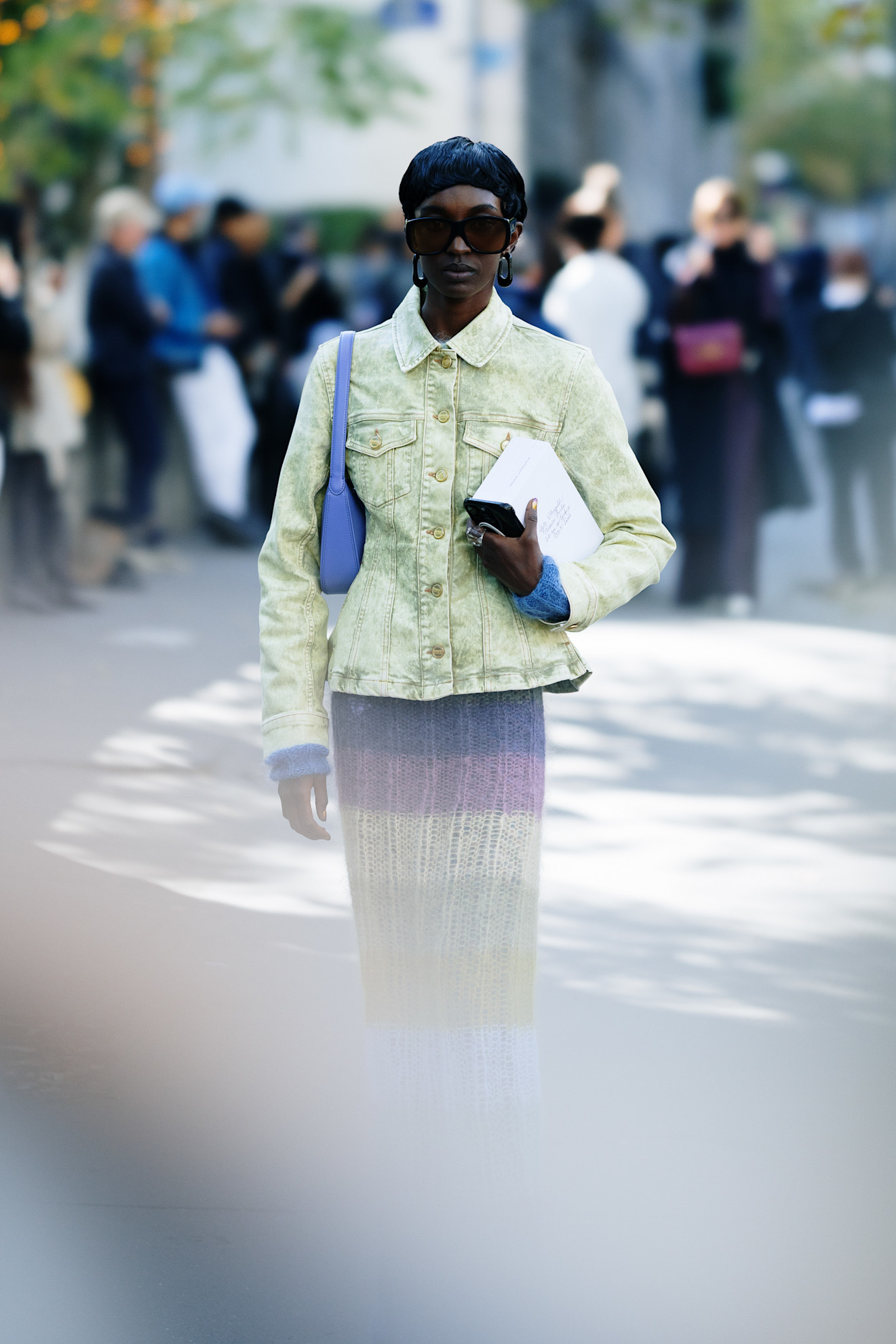 a woman wearing a light green denim jacket, blue bag and knitted skirt and a pixie cut facing the camera