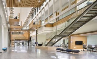 Inside The Taylor Institute of Teaching and Learning at the University of Calgary. Bright, large space, with white painted walls, and light wood elements. We see different spaces for conferences and learning to the right, through glass walls. A large, long staircase leads to the floors above.