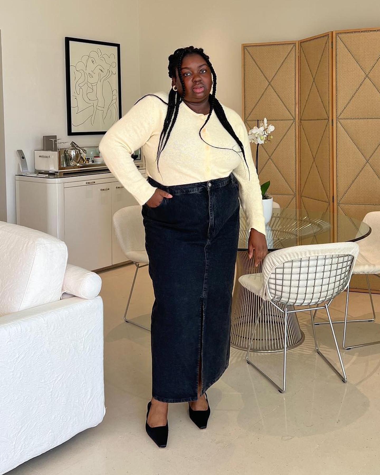 British plus size style influencer Abisola Omole poses in a pale yellow button-front cardigan, a dark-wash maxi denim skirt, and black suede slingback kitten heels.