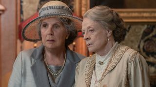 Penelope Wilton and Dame Maggie Smith stand together in conversation by the fireplace in Downton Abbey: A New Era.