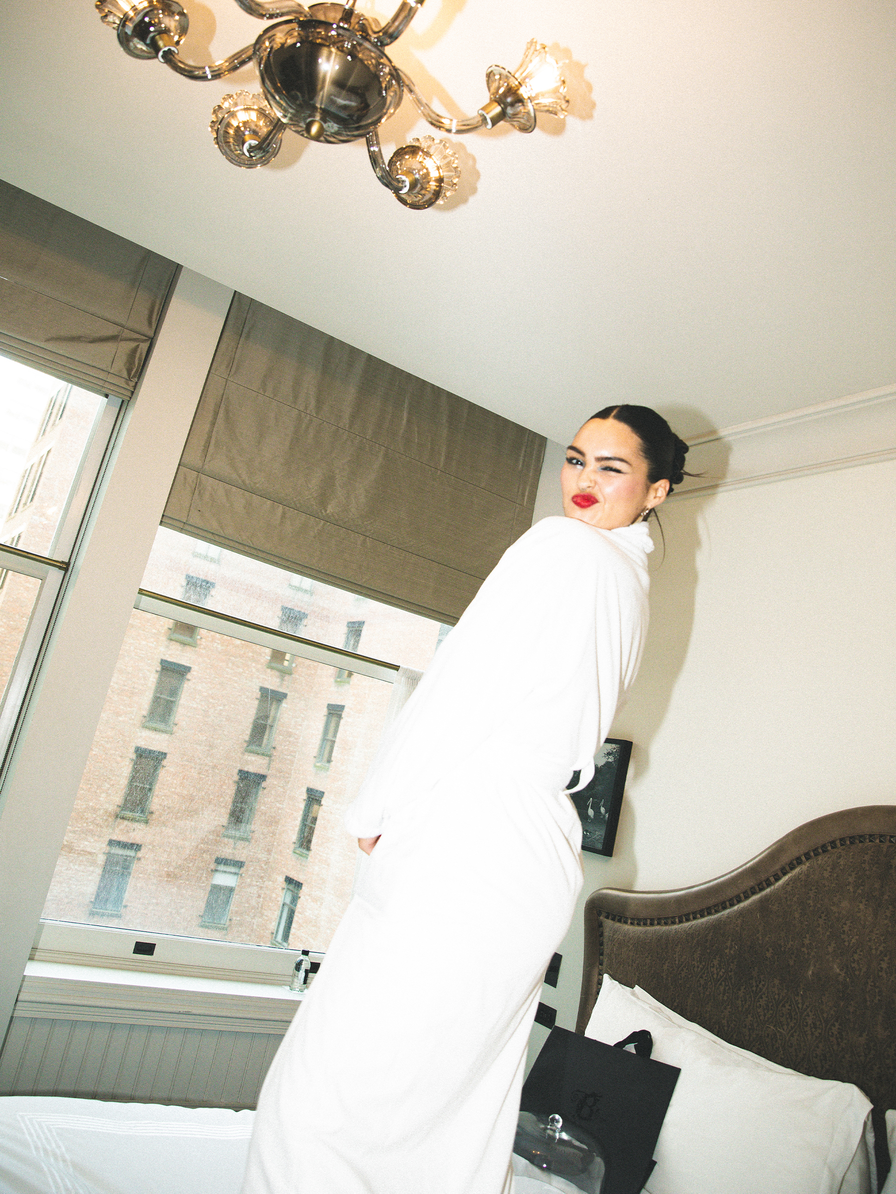 Anna Lambe poses in her hotel room. She is standing on the bed wearing a white robe and is winking at the camera.