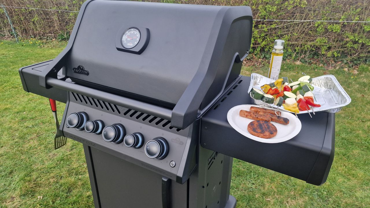 An example of how we test grills, The Napoleon Phantom Rogue on test, with a burger, some sausages, and vegetable kebabs resting on its side table