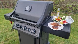 The Napoleon Phantom Rogue on test, with a burger, some sausages, and vegetable kebabs resting on its side table