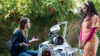 From left to right: Christa Miller talking with her hands while sitting at her rock table, while Jessica Williams stands and listens on the other side of the table.