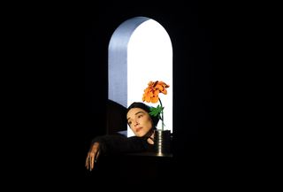 Annie Clark alias St. Vincent berpose melalui bingkai jendela hitam di samping bunga oranye besar.