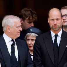 Prince William turns to Prince William and smiles at him, while both wear black suits with black ties and white shirts