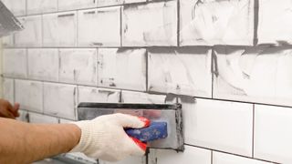 Gloved hand holding a grout float applying grey grout on white metro tiles