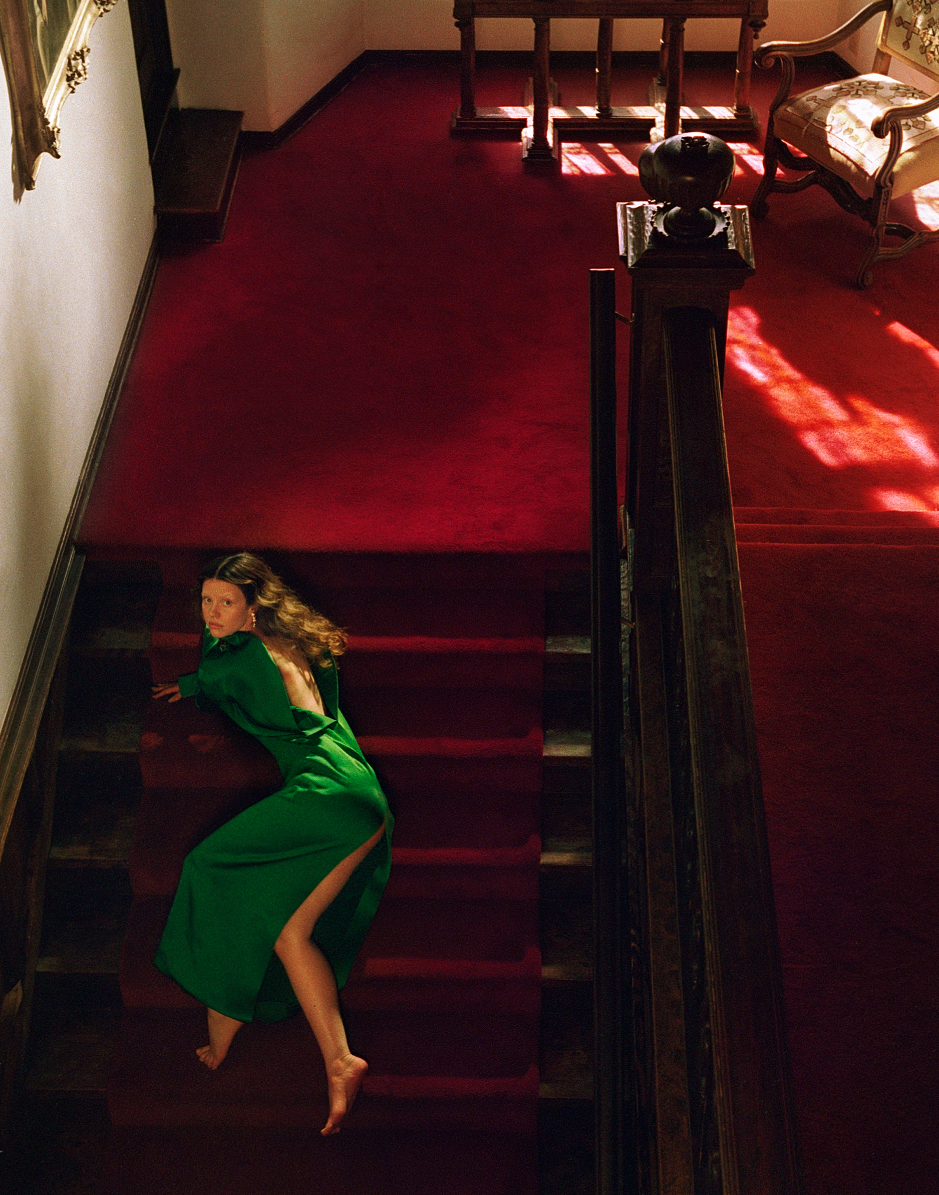 Image of Mia Goth from the October 2025 cover story. She is laying on a red carpeted stairs wearing an emerald green dress with open back.
