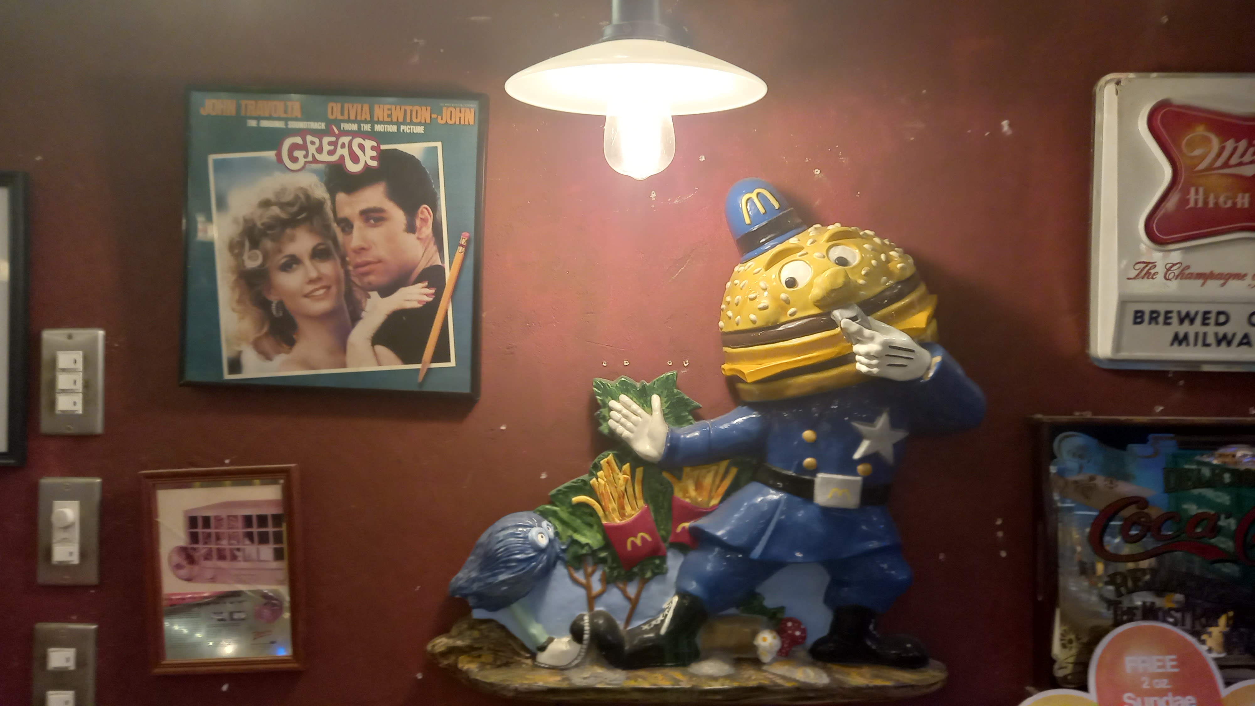 A vintage Mayor McCheese figurine in police uniform is displayed beneath a pendant lamp, surrounded by retro memorabilia including a Grease soundtrack poster and a Coca-Cola sign on a deep red wall.