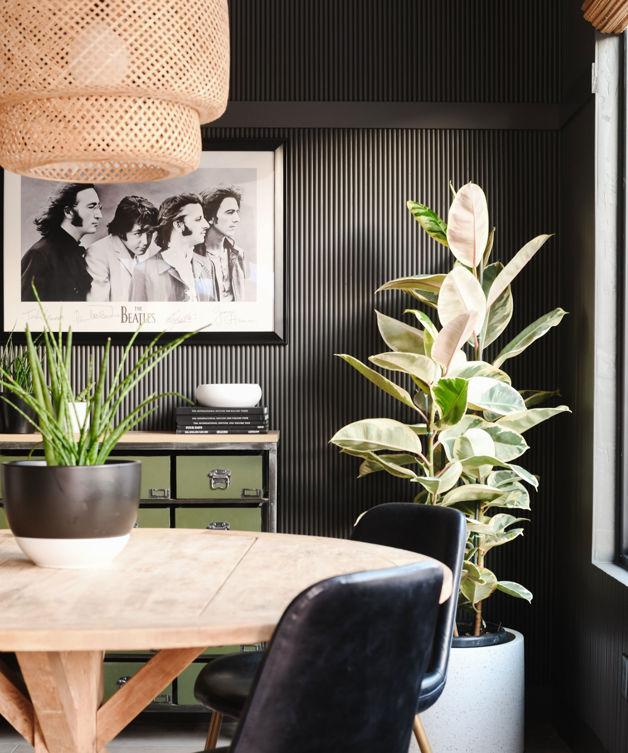 A living room diner painted in dark earthy gray-brown, with rattan light fitting, wooden table and pot plants.