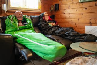 Senior couple in sleeping bags at home on sofa in livingroom smiling while watching tv although facing an energy crisis with rising electricity costs, they try to go through winter season with minimal heating, shallow focus on man