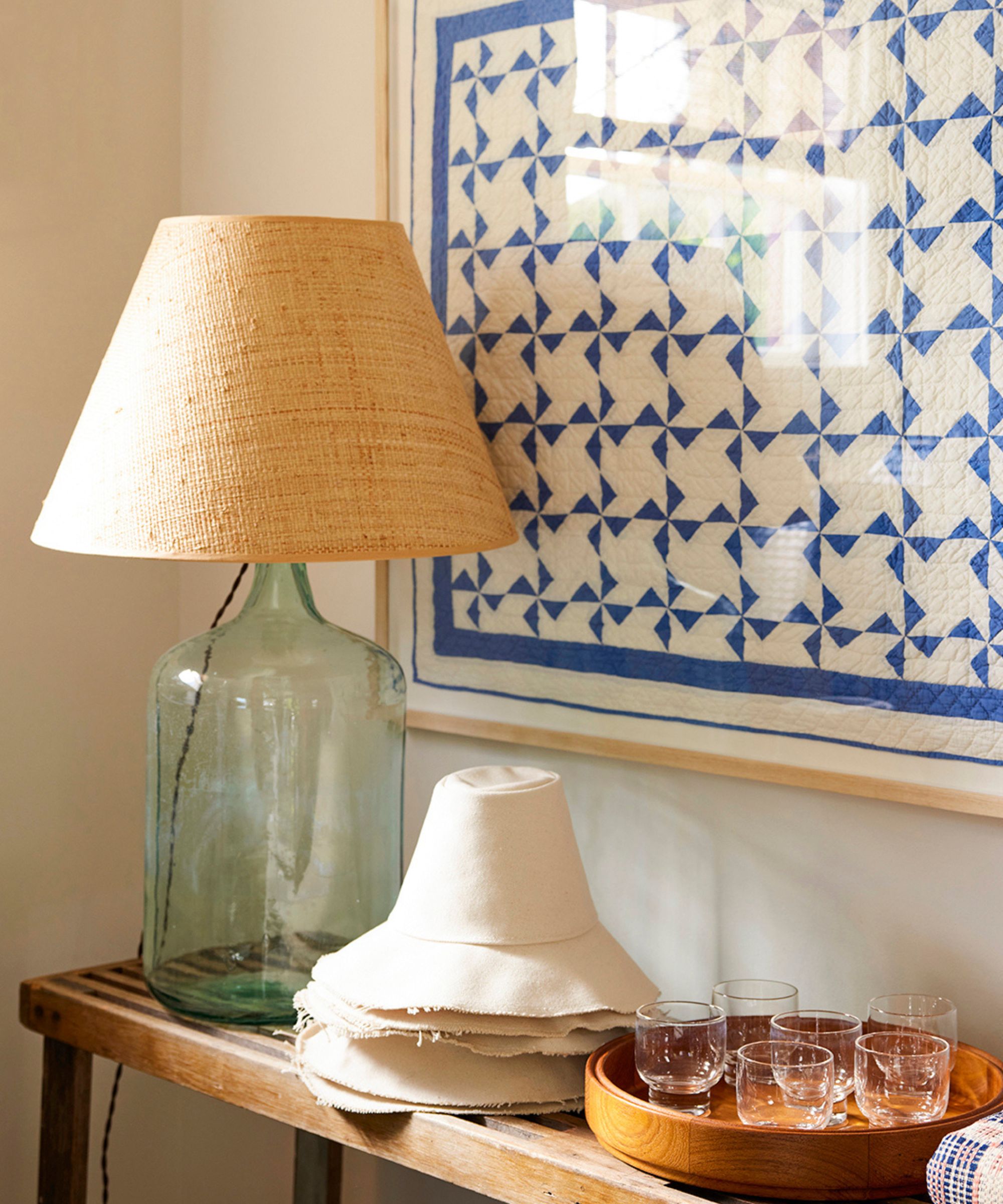 Close-up of a rustic wooden side table with a large glass lamp with a straw shade, stacked white hats, and a tray of small glasses, next to a framed quilt print.