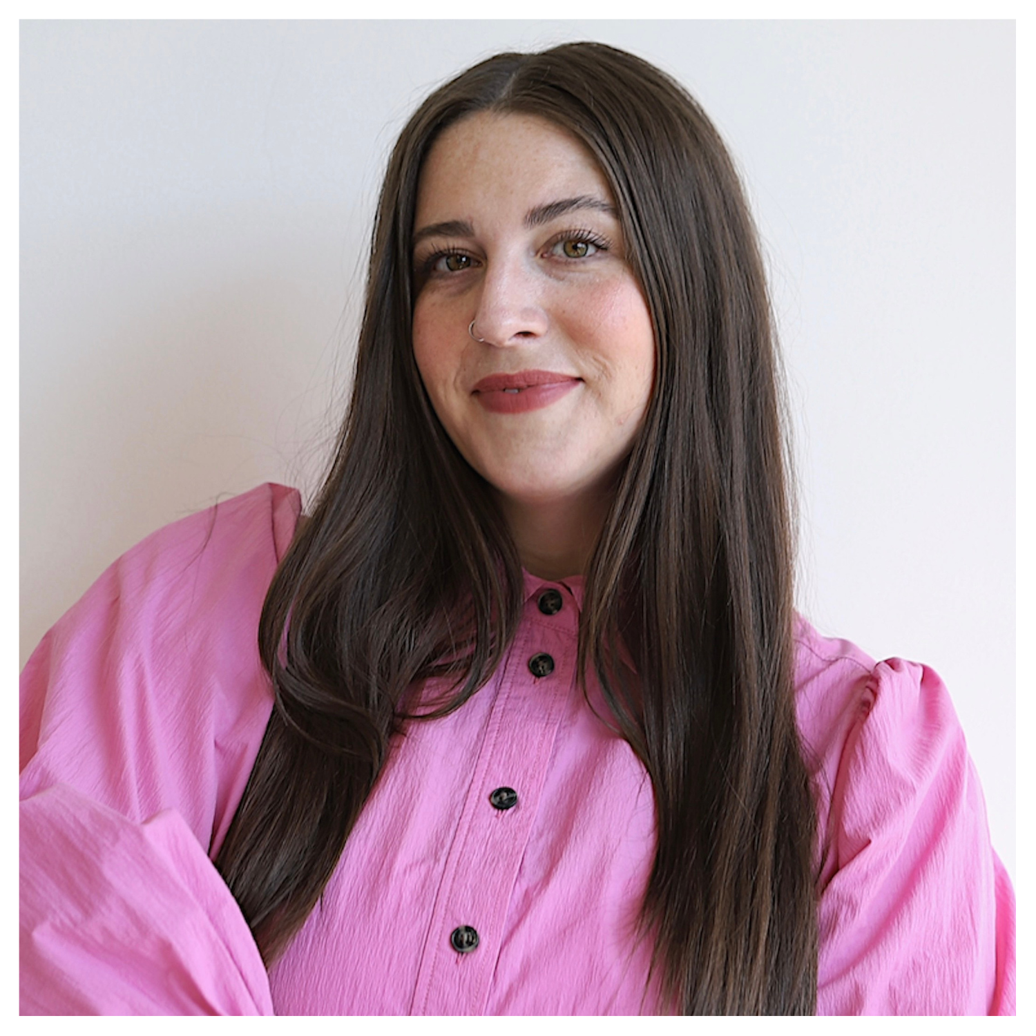 A lady with long, straight brown hair smiling in a puff-sleeved pink button down full sleeves shirt