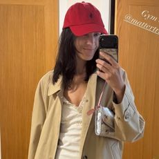 Alexa Chung takes a mirror selfie wearing a white cotton dress with a beige trench coat and a red cap.