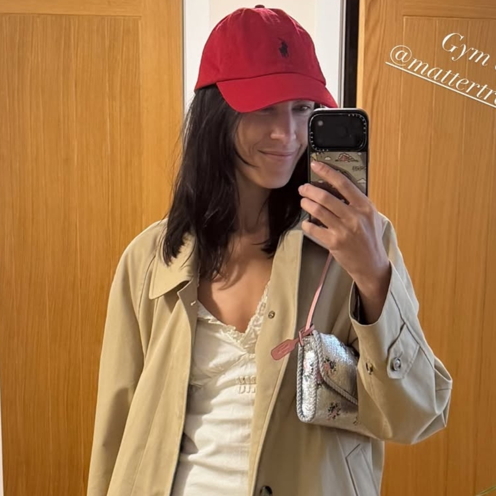Alexa Chung takes a mirror selfie wearing a white cotton dress with a beige trench coat and a red cap.