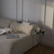 An image of a couch soaked in daylight with magazines, a laptop, and wine on top and beauty products on the side table.