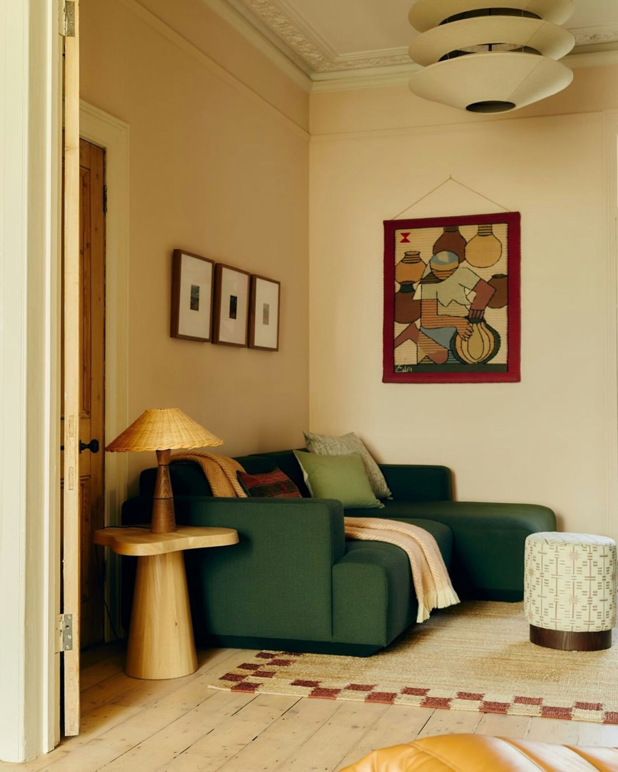 earth toned living room with green sofa, blush walls, blonde timber, a timber side table with lamp, crown molding, artwork on walls, and blush checkered rug on floor
