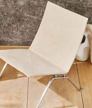 View of a light coloured, armless chair in a space with a dark patterned wall and wood flooring