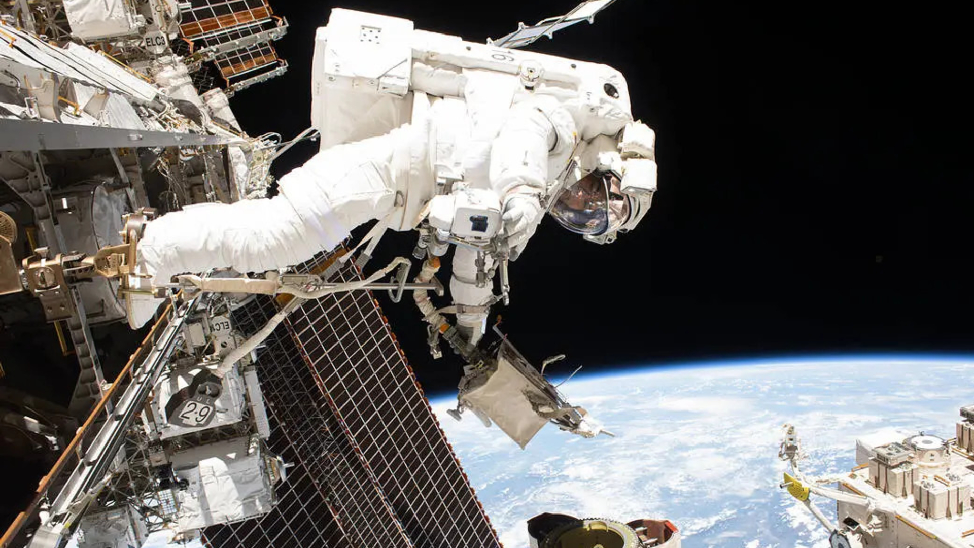 An astronaut in a white space suit faces toward the bottom of the image amidst a series of space equipment floating above Earth. 