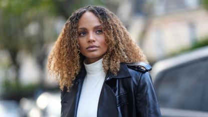 Alicia Aylies has curly hair and wears a white sleeveless / high neck body t-shirt, a black shiny leather varnished long trench coat, during a street style fashion photo session, on November 09, 2022 in Paris, France