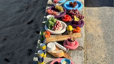 A series of colorful ceramic plates with a chunky design, sitting on a cement rim near a beach alongside cans of lemonade, baguette, and cheese, and filled with snacky bites.