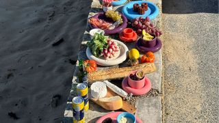 A series of colorful ceramic plates with a chunky design, sitting on a cement rim near a beach alongside cans of lemonade, baguette, and cheese, and filled with snacky bites.