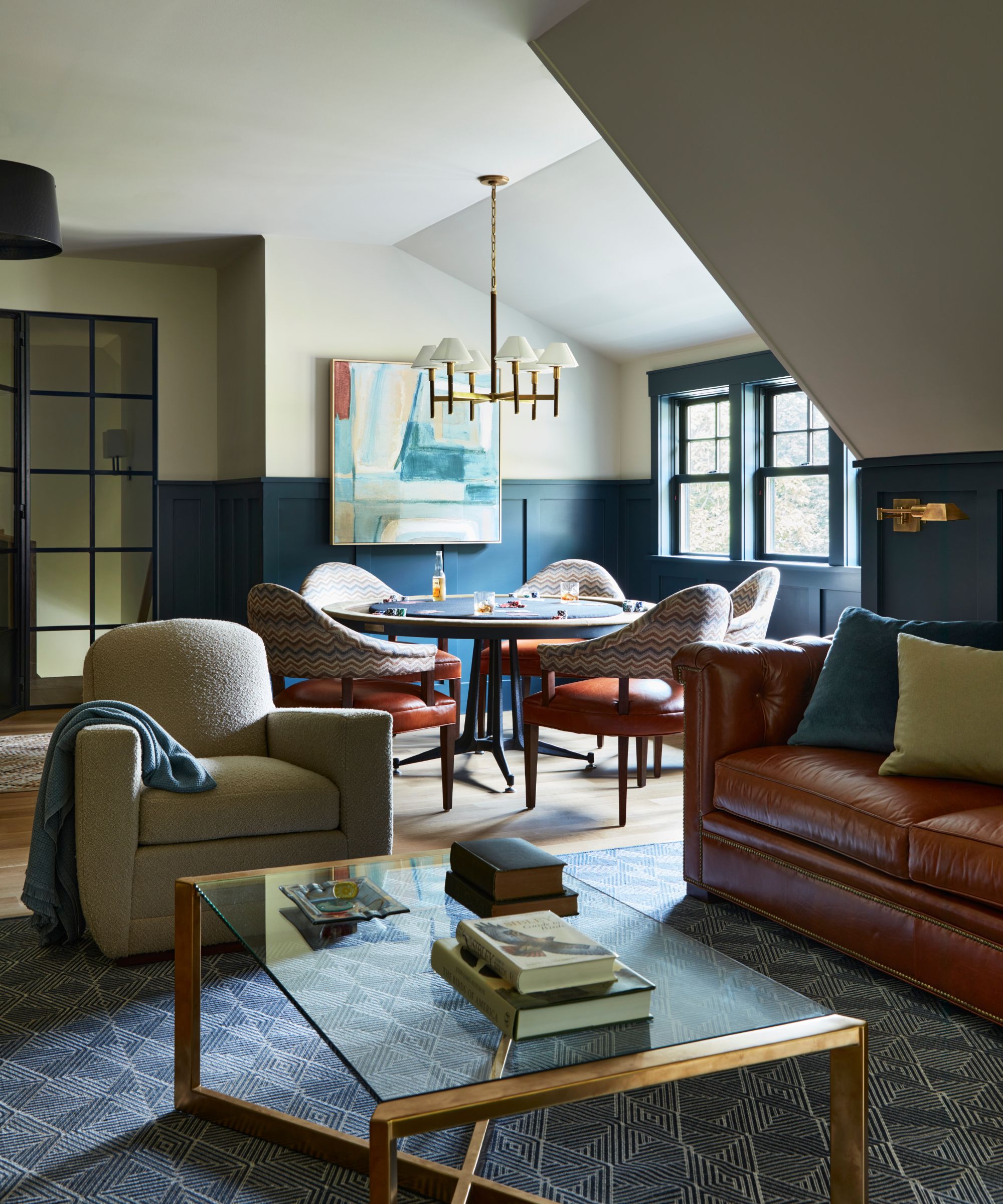 cozy blue home office designed by camden grace with navy two-tone panelled walls, a leather couch, armchair, gold and glass coffee table, and poker table in the corner