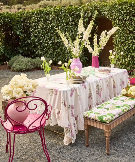 an outdoor tablescape with a floral tablecloth, a red metal chair, a bench with a floral cushion on it, and decorative flowers along the table