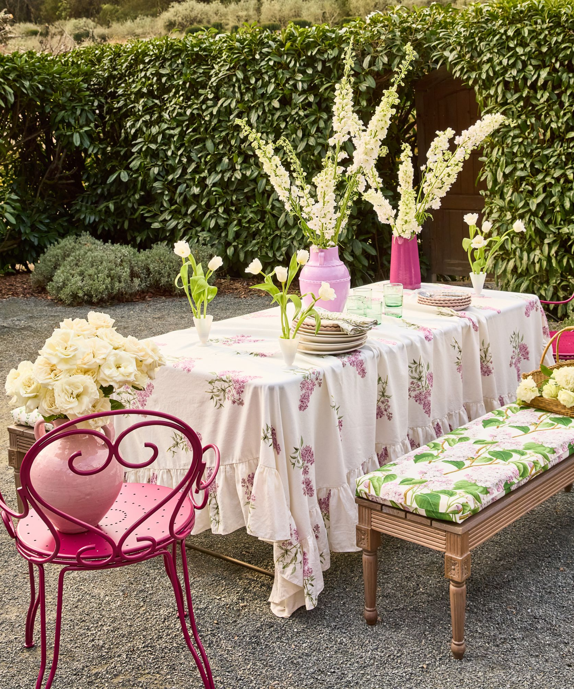 an outdoor tablescape with a floral tablecloth, a red metal chair, a bench with a floral cushion on it, and decorative flowers along the table