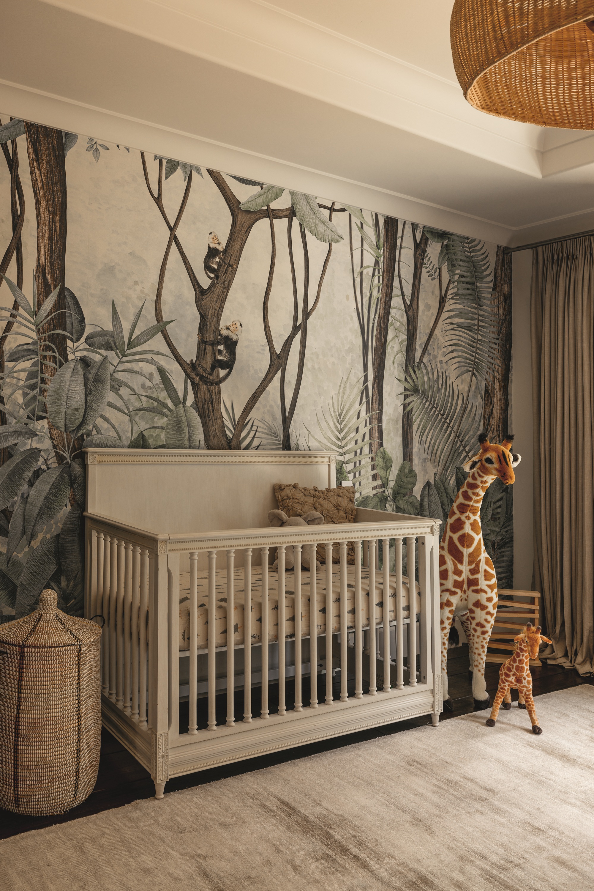 nursery with forest mural wallpaper, white ceiling, rattan pendant light, large giraffe toys, grey rug, rattan basket, white cot and heavy curtains