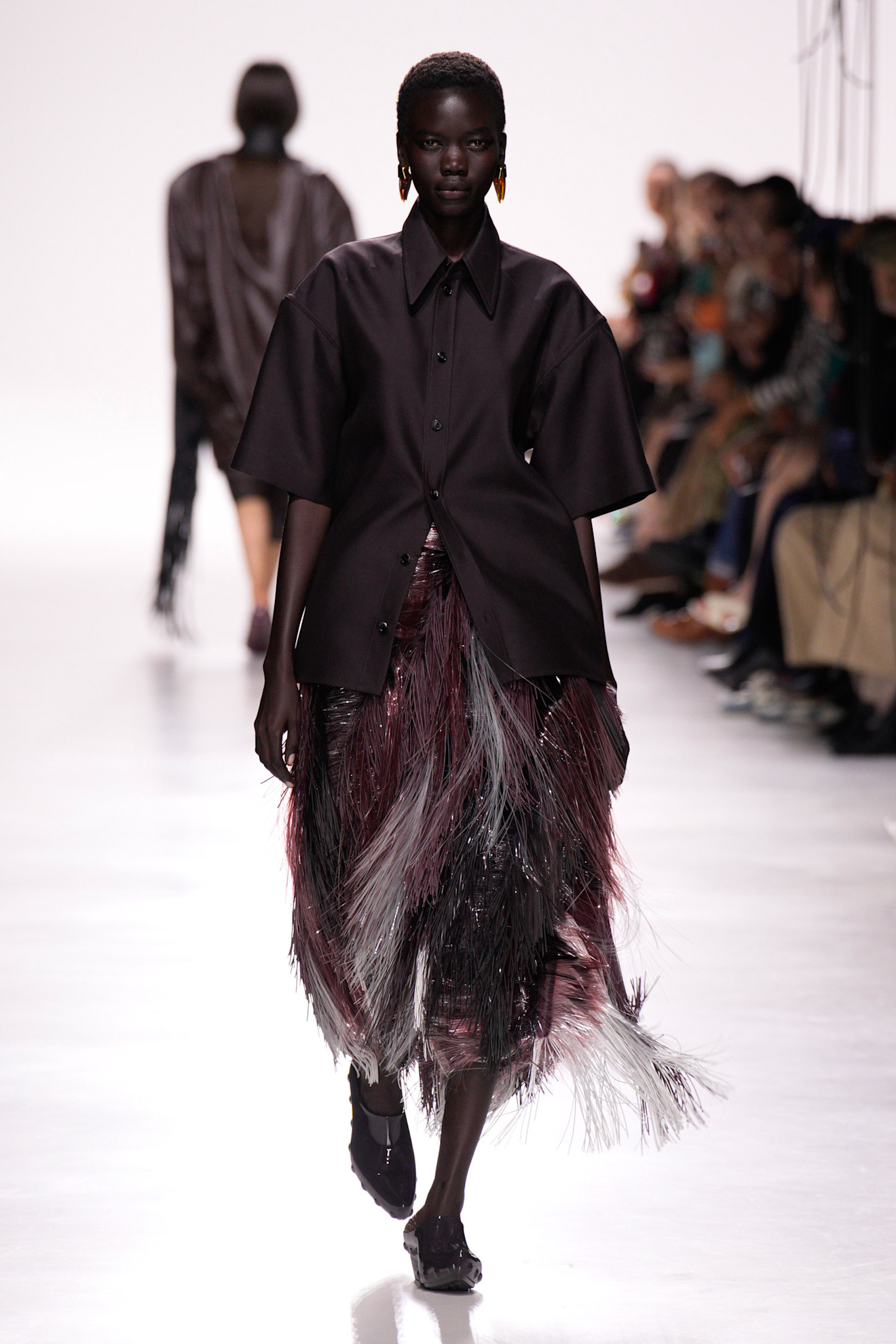 A model at the Bottega Veneta S/S 26 show wearing a button-down short-sleeve shirt and a fringe skirt with clogs.